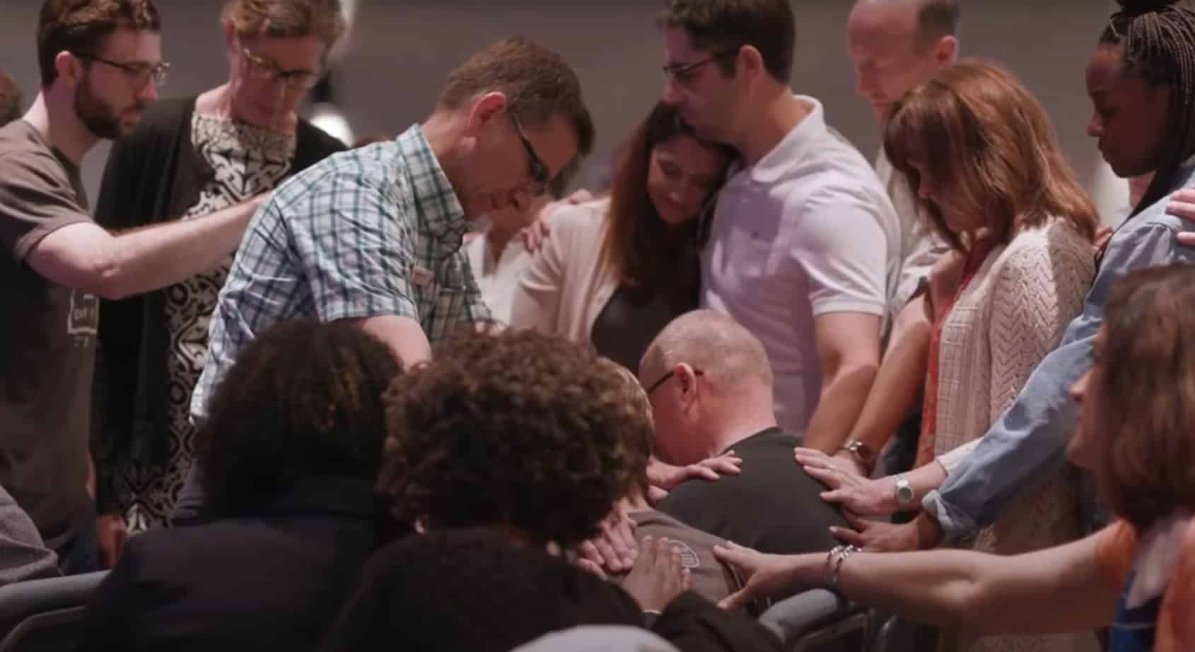 A group of people stand closely together, placing their hands on a seated man's shoulders in a supportive gesture.